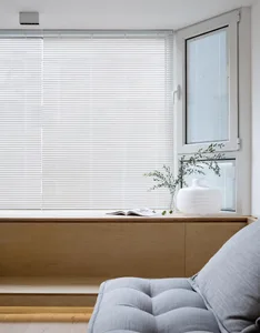 A minimalist living room featuring a wooden window sill with a plant and a vase on it, a grey tufted sofa, and sunlight strea