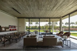 Modern living room with concrete walls, large windows overlooking a pool and garden, and a comfortable sofa and dining table.