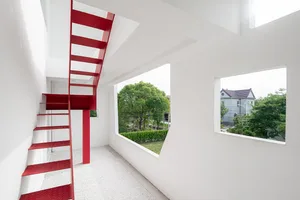 A minimalist, white interior with a striking red metal staircase and large windows.