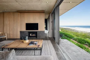 A modern living room with wooden furniture, large windows overlooking the sea, and a fireplace.
