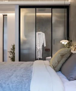 Bedroom featuring a white bed with a beige pillow, a glass sliding door with a white robe, and a plant in the corner.
