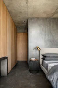 Minimalist bedroom with concrete walls, wooden furniture, and a bed with grey bedding.