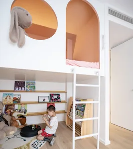 A modern children's room with a white loft bed, bookshelves, and a child playing on the floor with toys and books.