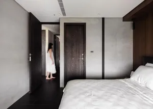 A minimalist bedroom with concrete walls, a white bed, and a person walking through the hallway.