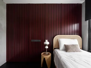 Modern, minimalist bedroom with a bed, bedside table, and lamp against a dark red wall.