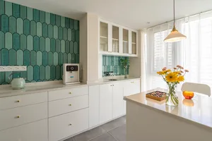 A modern kitchen with white cabinets, ceramic green tiles on the wall, white countertops, and a white chair.