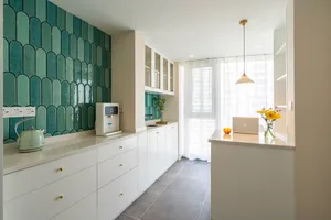 A modern, minimalist kitchen featuring green ceramic tiles, white cabinetry, and a white countertop with a kettle, laptop, an