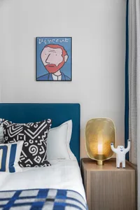 A modern bedroom featuring a blue upholstered headboard with patterned throw pillows and a golden lamp on a side table.
