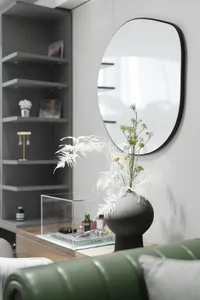 A modern home office with concrete and glass elements featuring a green sofa, a round mirror, a black vase with dried plants,