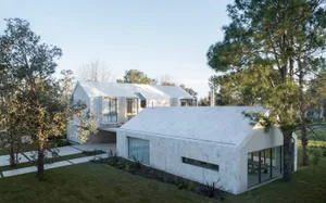 Modern, minimalist residential exterior with white concrete walls and large windows, surrounded by greenery.