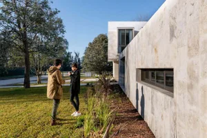 A modern house with a concrete exterior featuring a large window and greenery around it.