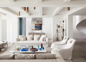 A modern white living room with a white sofa, armchairs, and coffee table with books and glass vases.