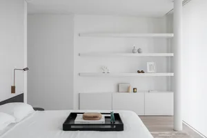 A modern minimalist bedroom featuring a white bed with wood headboard, steel-and-white shelving unit, and white walls.