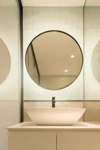 A modern bathroom features a circular mirror hanging above a white sink, surrounded by ceramic tiles with a sleek, minimalist