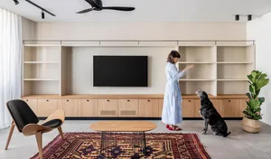 A modern living room with a wooden coffee table, a black armchair, and a woman arranging items on a white shelving unit.