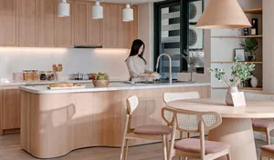 A modern kitchen with light-colored wood cabinets and a matching breakfast bar, featuring a woman washing dishes at the sink.