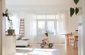 A minimalist living room featuring white window curtains, hardwood floors, and a wooden ladder.