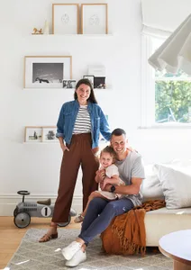 A family with one child sitting on a couch in a living room with neutral-toned decor and framed art on the wall.