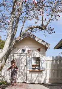 A small, minimalist children's playhouse made of wood with a door adorned with colorful triangular flags and a white picket f