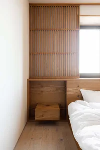 A modern, minimalist bedroom with wooden storage units, white bedding, and a window.