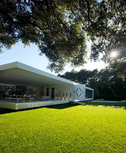 Modern white house with a concrete exterior and large glass windows, surrounded by green grass and trees.