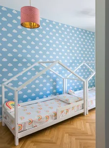 A playful kids' bedroom featuring a white bed frame resembling a house, rainbow-patterned sheets, and cloud-patterned wallpap