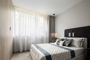 A modern minimalist bedroom with white curtains, wood flooring, and a double bed with geometric bedding.