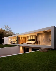 A modern residential exterior with concrete walls and a glass front door, illuminated by warm indoor lighting against a clear