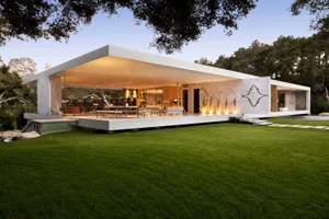 A modern minimalist residential exterior with glass walls and a flat roof, surrounded by a lush green lawn.