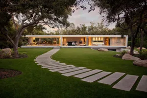 A modern residential home with a concrete pathway leading up to the entrance, surrounded by green grass, rocks, and trees.