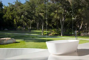 Modern, minimalist outdoor bathroom featuring a concrete bathtub and polished floor, surrounded by lush greenery and trees.