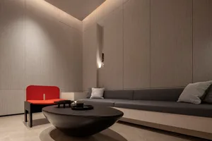 A minimalist living room with a curved concrete coffee table, a red accent chair, and a built-in gray sofa with patterned thr