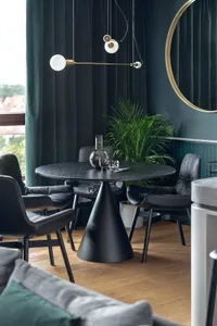 A modern dining room featuring a round concrete table surrounded by black leather chairs, with green curtains and a potted pl