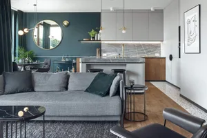A modern living room with grey sofa, black coffee tables, green cushions, and a kitchen counter with a barstool.