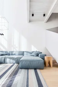 A modern minimalist living room featuring a fabric sofa, wooden stool, and a striped rug.