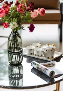 A modern interior featuring a glass coffee table with a vase of vibrant flowers, a teapot and cups, and a rolled-up magazine.