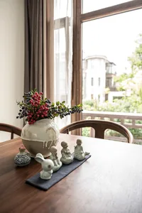 A wooden table with a ceramic vase filled with berries and small ceramic figures on a blue mat, set against a backdrop window