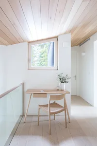 A minimalist home office with a wooden desk and chair, featuring a small window and natural lighting.