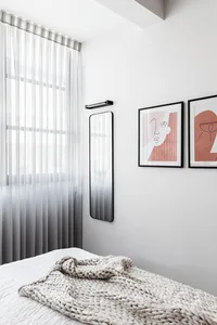 A minimalist bedroom featuring a white bed with a textured blanket, framed artworks, and sheer curtains allowing natural ligh
