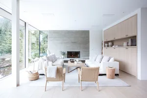 A modern minimalist living room with white furniture, a wooden floor, and a large glass wall overlooking a forest.