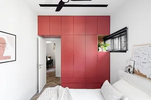 A modern minimalist bedroom featuring a large red cabinet, white bed, and white walls, with a view into a hallway with herrin