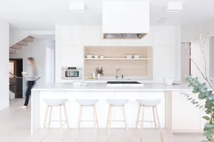 A modern minimalist kitchen featuring a white island with four stools, light-colored wood cabinets, and stainless steel appli
