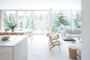 Modern minimalist living room interior with white walls, hardwood floors, and large windows showcasing snowy trees outside.