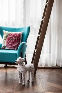 Teal upholstered armchair with a floral cushion, standing on a wooden floor with white sheer curtains.