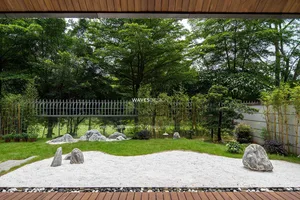 A minimalist outdoor garden featuring a dry landscape of rocks and sand, surrounded by lush greenery and a wooden fence.