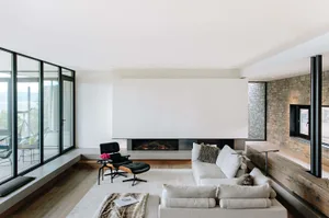 Modern living room with a fireplace, a white sectional sofa, and a black armchair with a footstool. The room features a stone