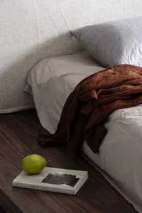 A cozy minimalist bedroom with a bed covered in white linen and a brown knit sweater draped over it, a green pear resting on 