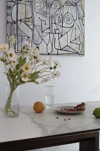 A vase of pink and white flowers, a glass of water, a plate of pomegranate seeds, and a piece of green fruit on a marble tabl