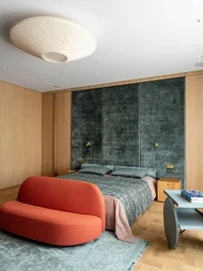 A modern bedroom featuring a velvet bed against a wooden accent wall, with a red sofa and a blue coffee table.