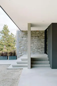 Modern front porch featuring a concrete entrance with steps, a stone wall, and a tree in the background.